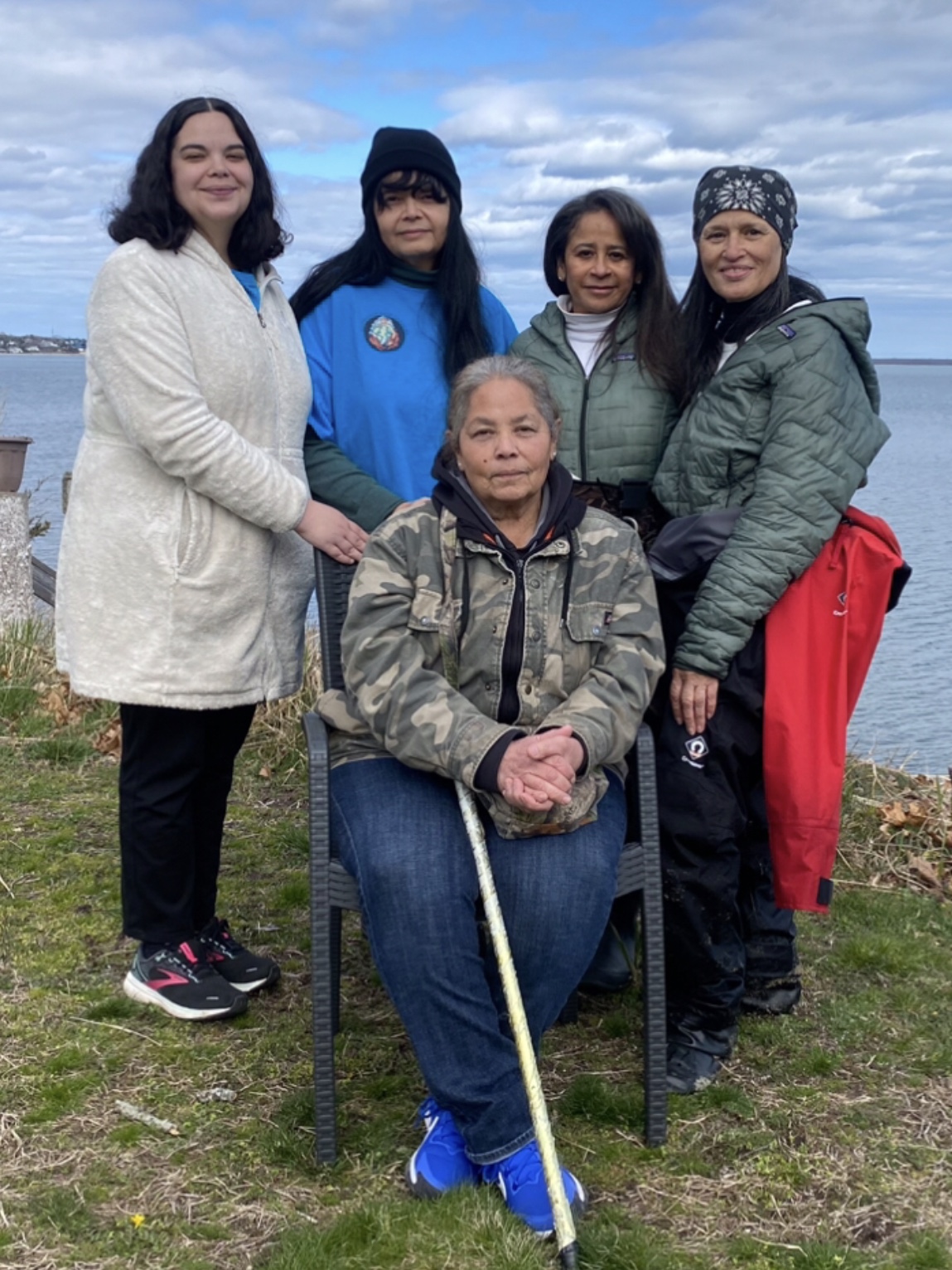 Five Shinnecock Farmers posing together outdoors, with a serene lake and cloudy sky in the background. One person is seated in the front, holding a cane, while four others stand behind, smiling.
