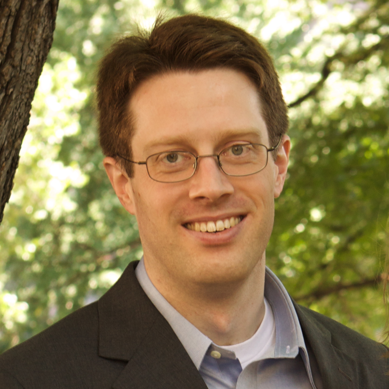 Portrait of a smiling person wearing glasses and a dark blazer over a blue shirt, standing outdoors with trees in the background.