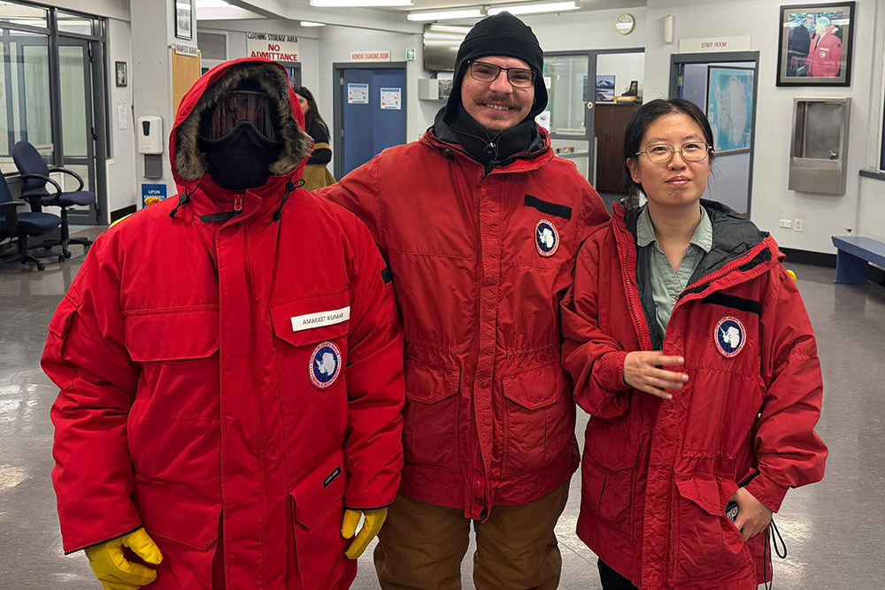Geosciences graduate students Three people in red Antarctic parkas stand indoors. One is fully covered, the others smile. The scene is warm, despite the heavy gear.