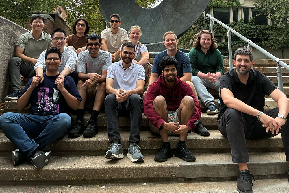 Jesus Perez Rios and Research team  Group of eleven individuals From The Stony Brook University Department of Physics and Astronomy posing on steps outdoors, smiling, with a large sculpture in the background.