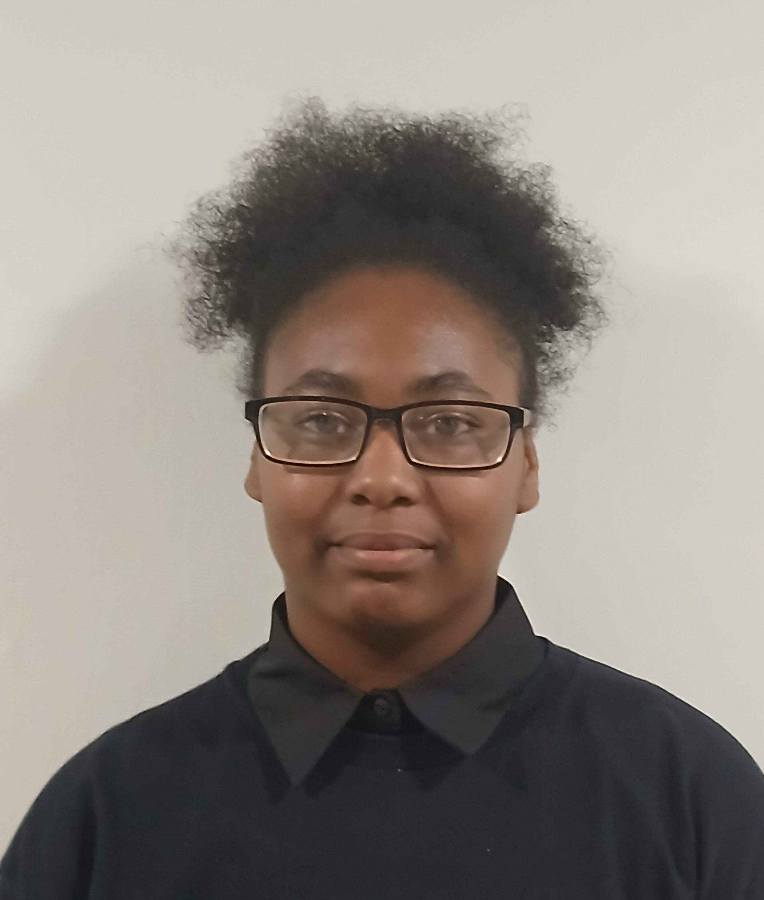 A person with curly hair and glasses wears a black collared shirt. They stand against a plain white background, conveying a neutral expression.