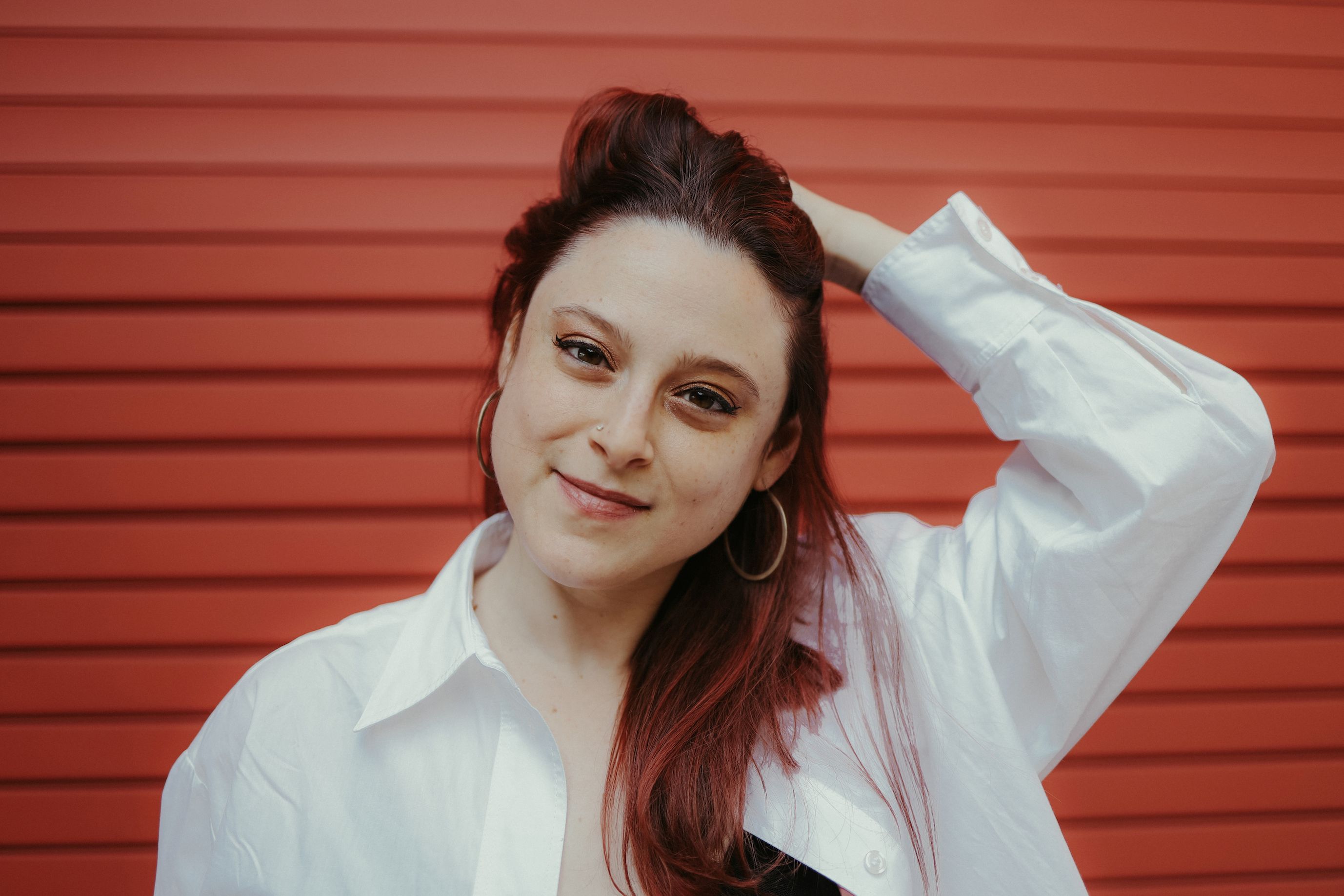 Stony Brook University Art History Phd Candidate posing in front of a red background wearing hoop earrings and white button down shirt with her left hand in her hair.
