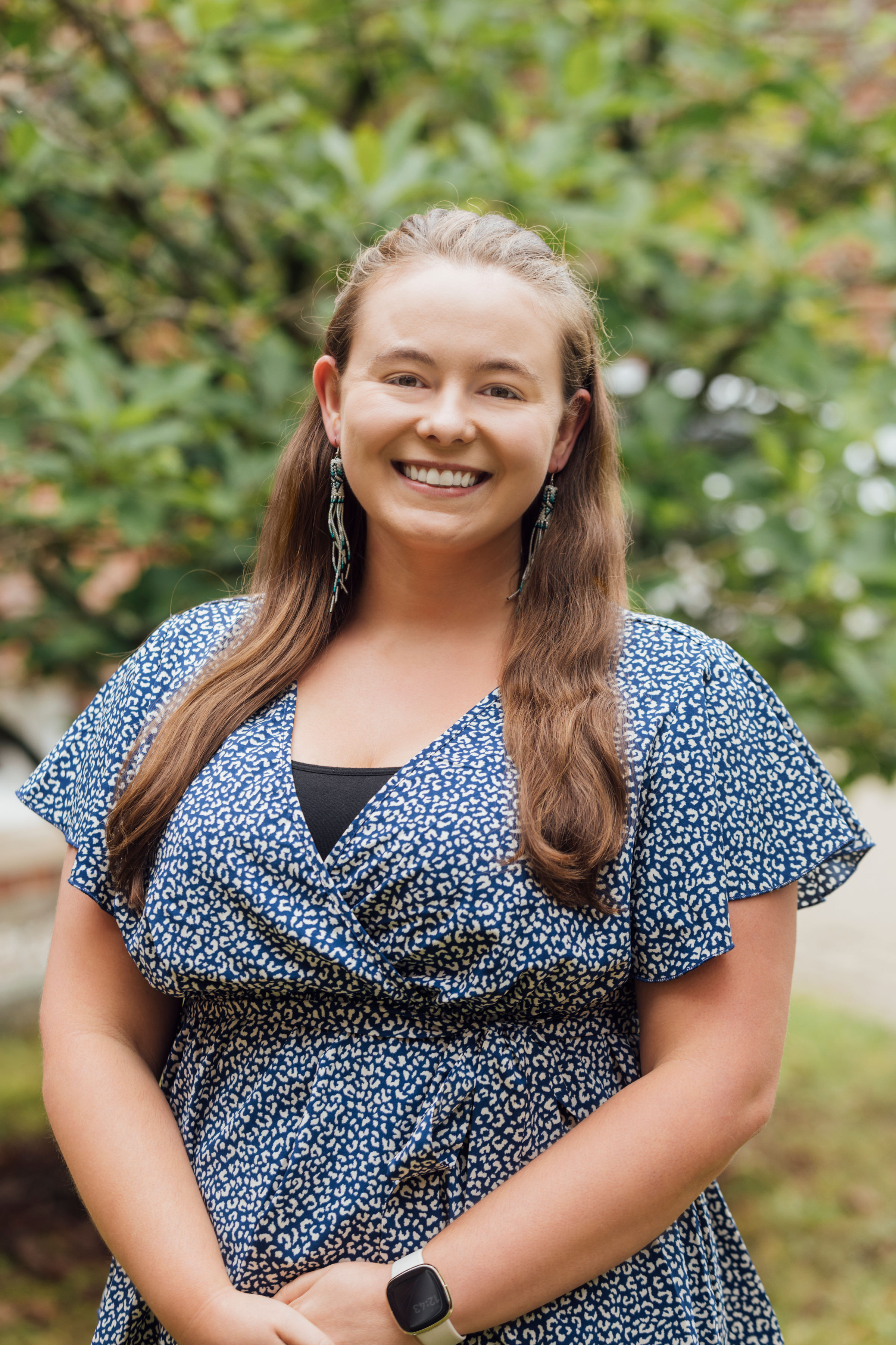 Darcey Evans A person smiling at the camera, wearing a blue patterned dress, standing in front of a leafy background.