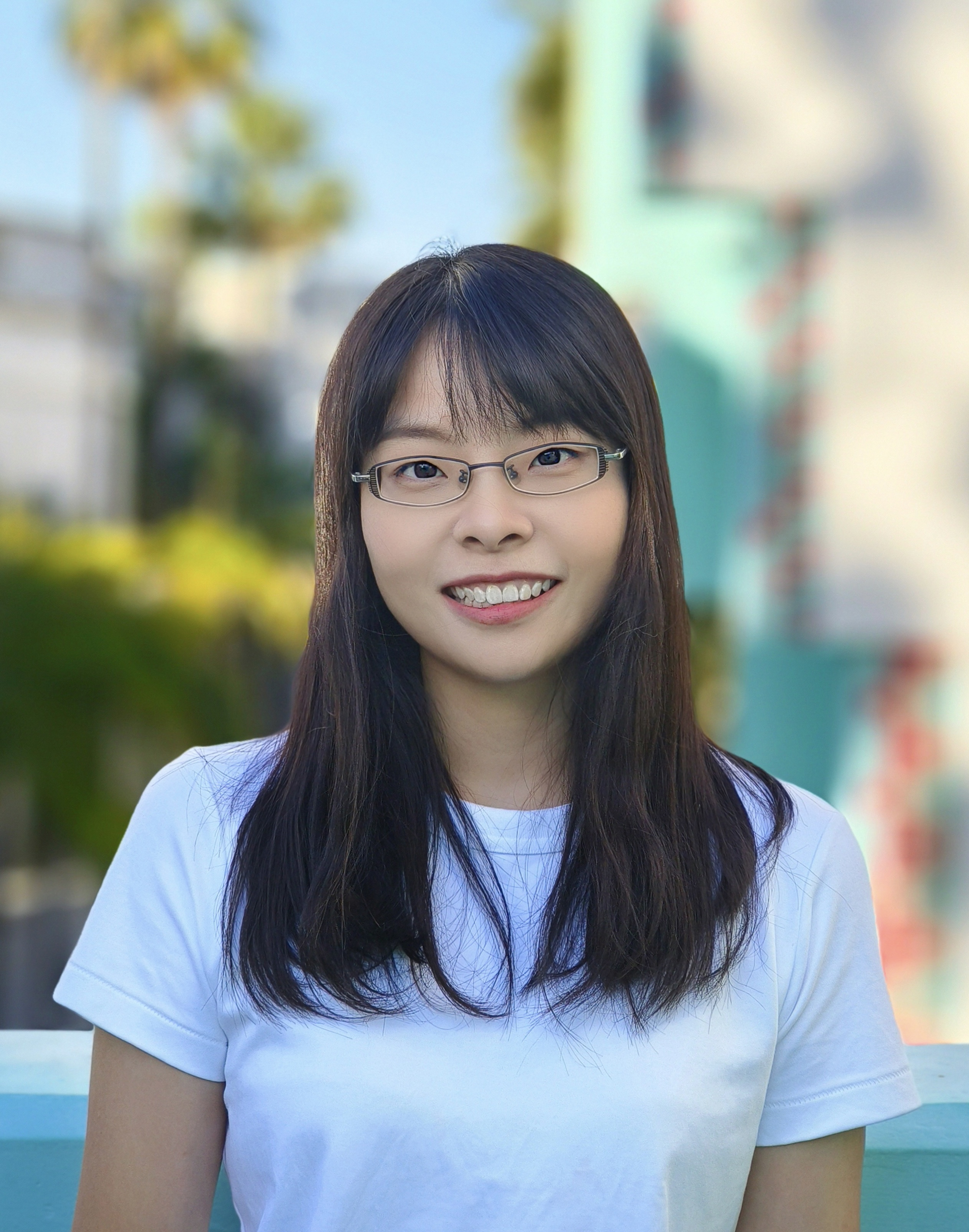 Hui Wang Portrait of a smiling person wearing glasses and a white t-shirt, standing outdoors with blurred trees and a blue sky in the background.