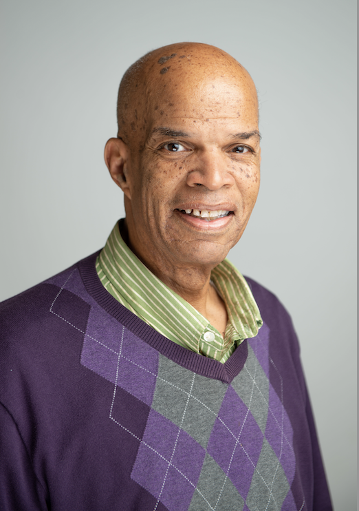 Mark Chambers Portrait of a smiling individual wearing a purple and gray argyle sweater over a green striped shirt, against a light gray background.