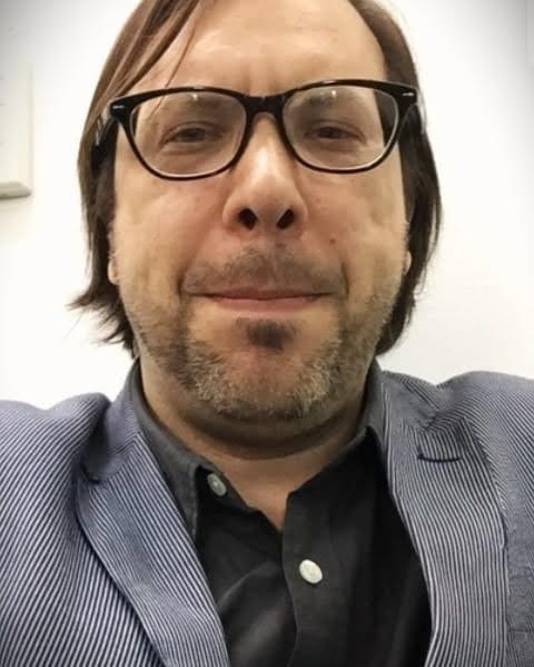 Michael Loncar Close-up portrait of a person wearing glasses and a blue jacket over a dark shirt, smiling slightly at the camera.