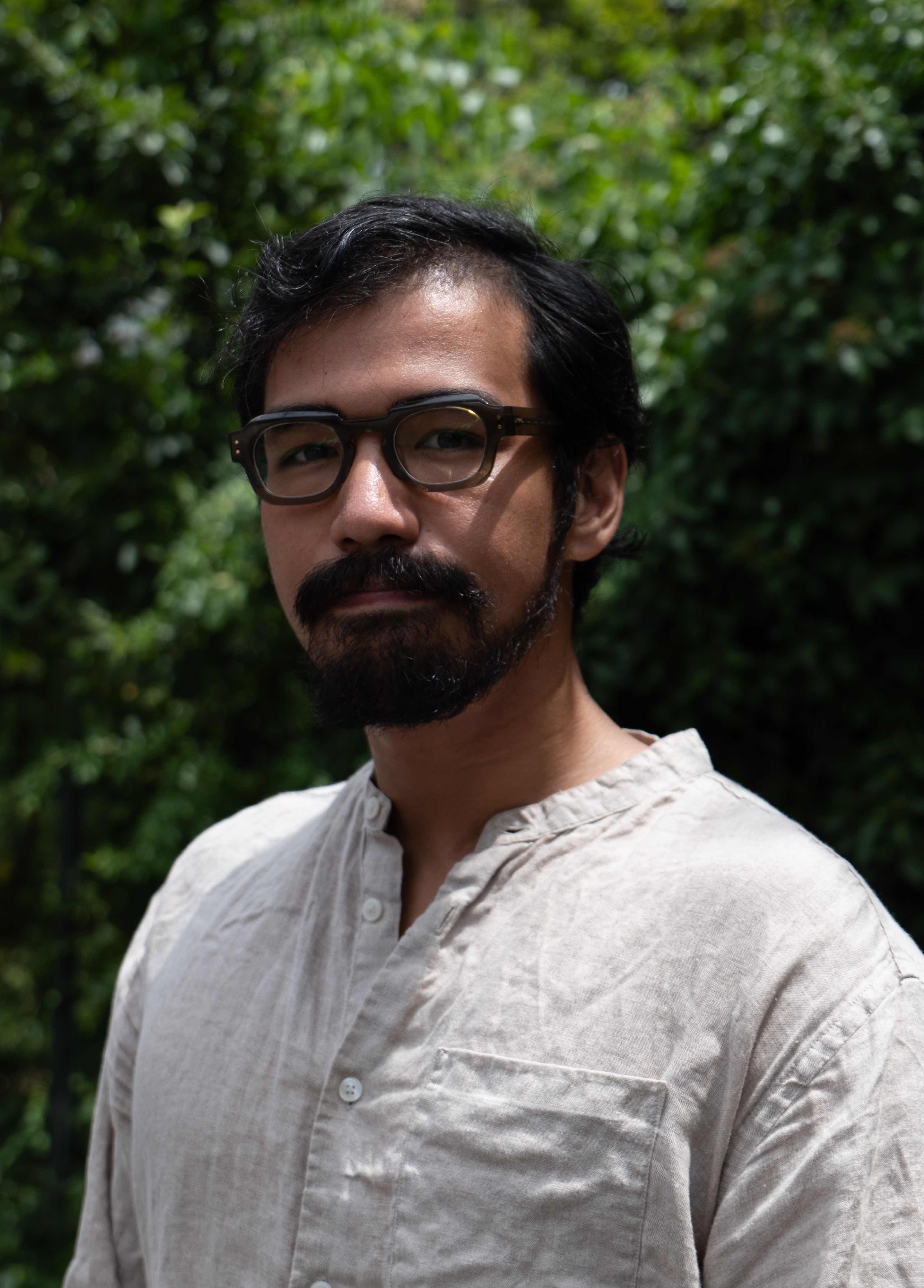 Riga Shakya Person wearing glasses and a light beige shirt standing in front of a green, leafy background.