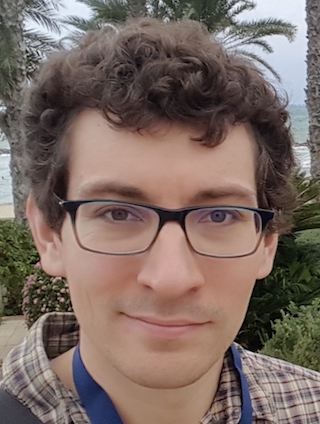 Ross Corliss Person wearing glasses and a plaid shirt standing in front of a brick wall with vines.