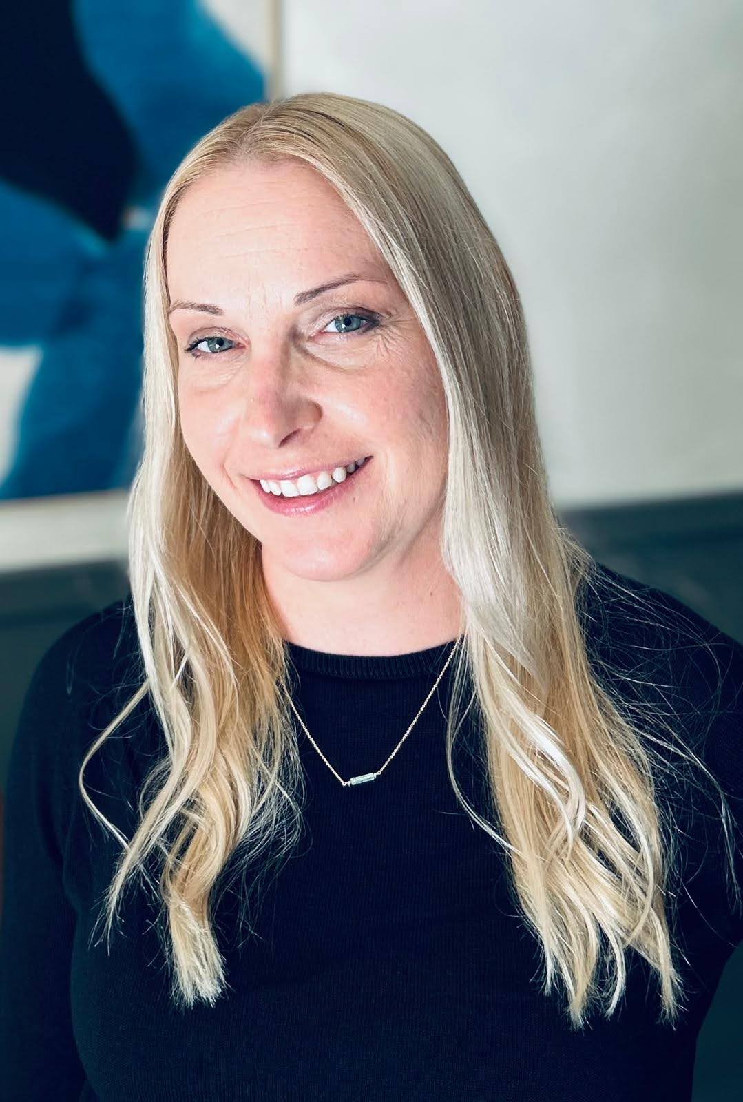 Serena Clark  Person with long blonde hair smiling in front of a blue abstract art piece.