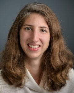 Kristie Schlauraff Portrait of a smiling person with long, wavy hair, wearing a light-colored scarf over a blouse against a solid dark background.