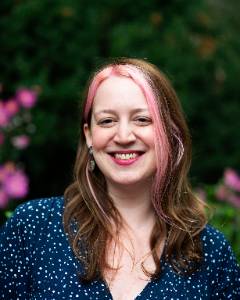 Alison Fairbrother Person smiling in a blue polka dot top, with pink highlights in their hair, standing in front of green foliage with purple flowers.