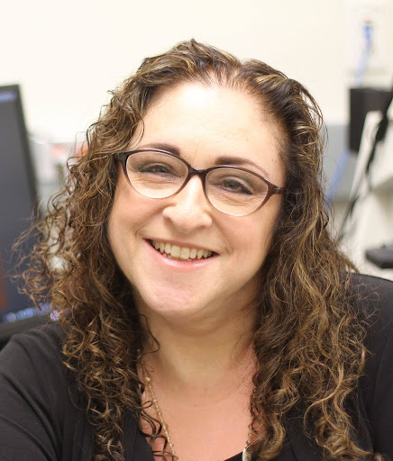 Amalia Napoli A smiling person wearing glasses and with curly hair, seated indoors in an office setting.