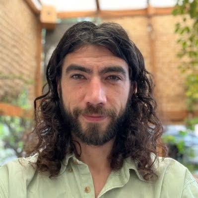 Cameron Jones Portrait of a smiling person with long curly hair, wearing a light green shirt, outdoors.