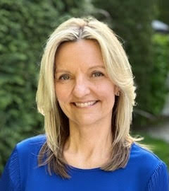 Donna Downing Portrait of an individual smiling, wearing a blue top, set against a blurred green background.
