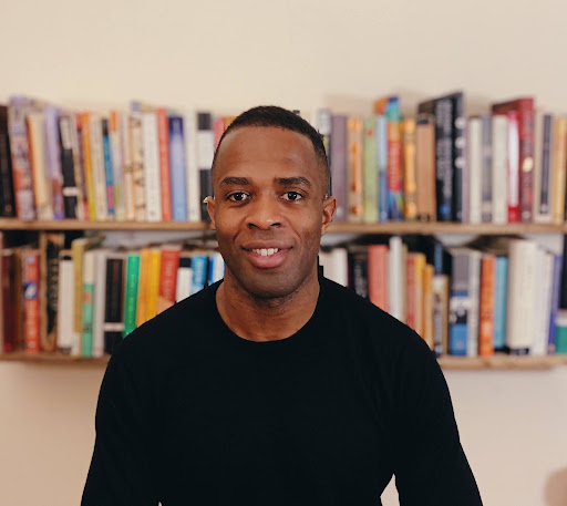 Eskor David Johnson Person in black shirt smiling in front of a bookshelf filled with colorful books.