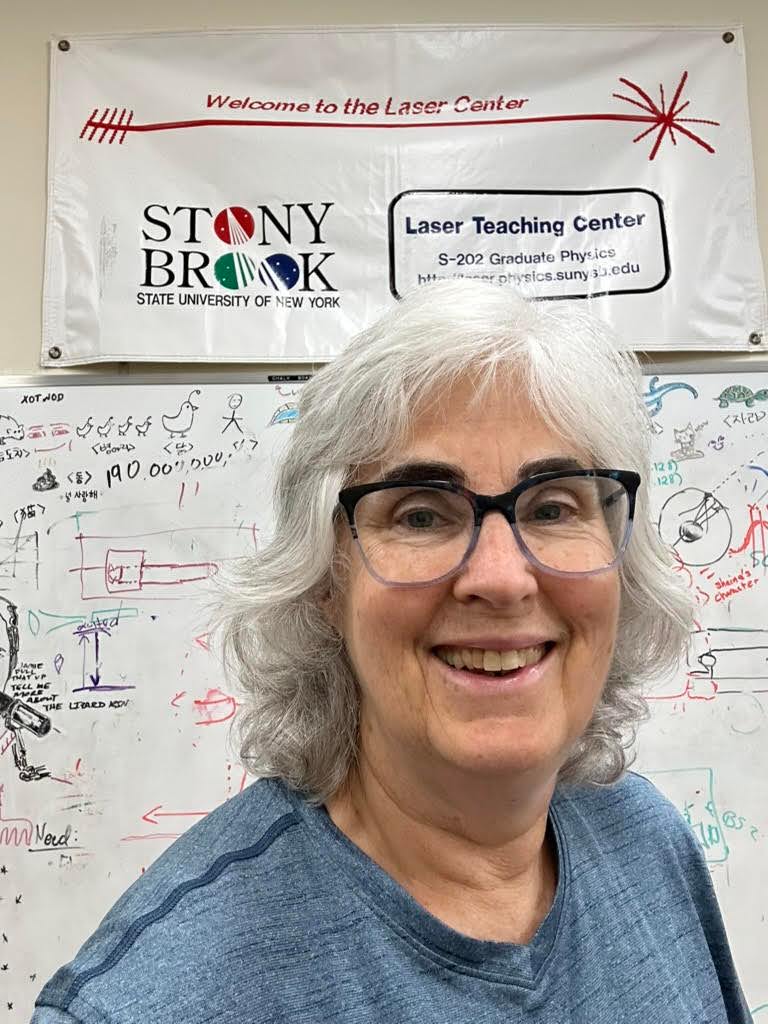 Gillian Michaels Individual smiling in front of a 'Welcome to the Laser Center' sign at Stony Brook University, with a whiteboard covered in colorful mathematical equations in the background.