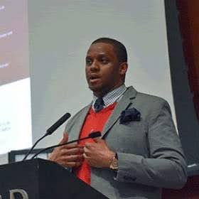 Jarrett Drake A person speaking at a lectern with a microphone, wearing a gray suit and red sweater.