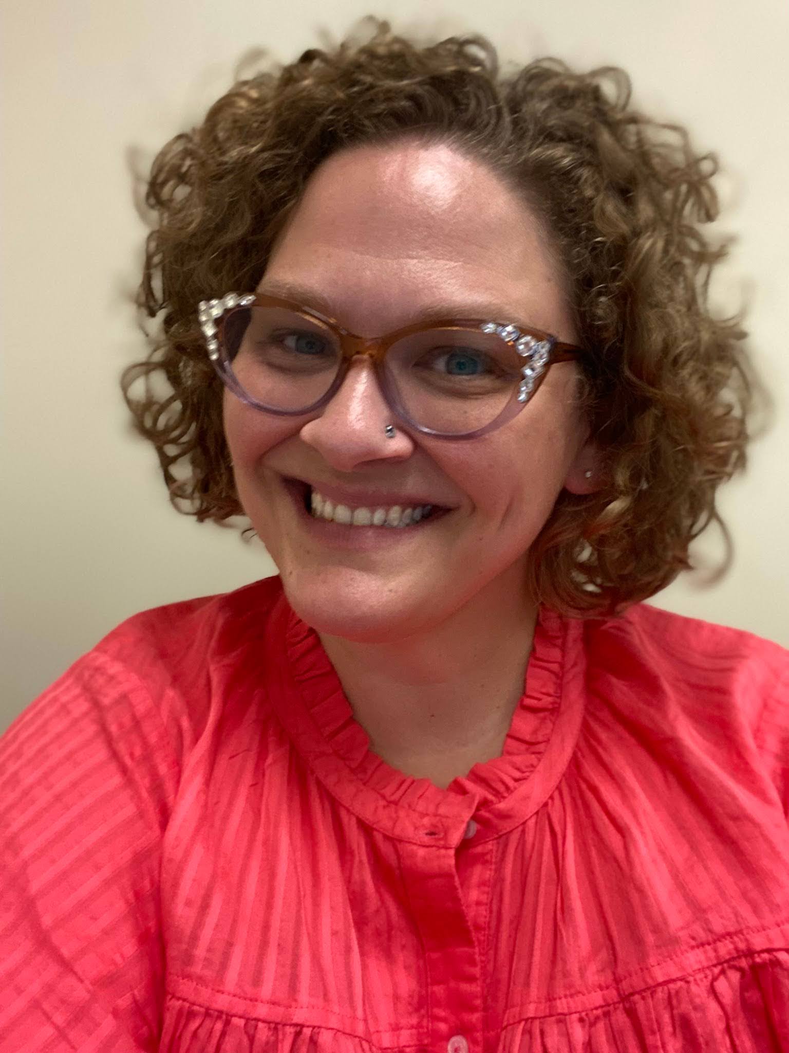 Jennifer McConnel Person with curly hair and glasses smiling in a red shirt.