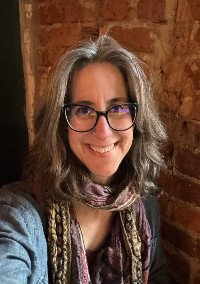 Jordanna Sprayberry Person with glasses smiling at the camera, wearing a scarf and a patterned jacket, standing in front of a brick wall.