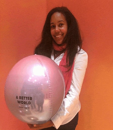 Karina Beras A person smiling and holding a large balloon with the phrase "A Better World" printed on it, against a vibrant orange background.
