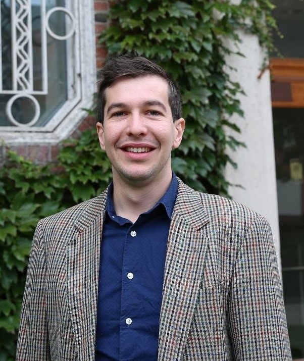 Lorenz Ekerdt A person smiling, standing outside a building covered with ivy. The person is wearing a plaid blazer over a blue shirt.