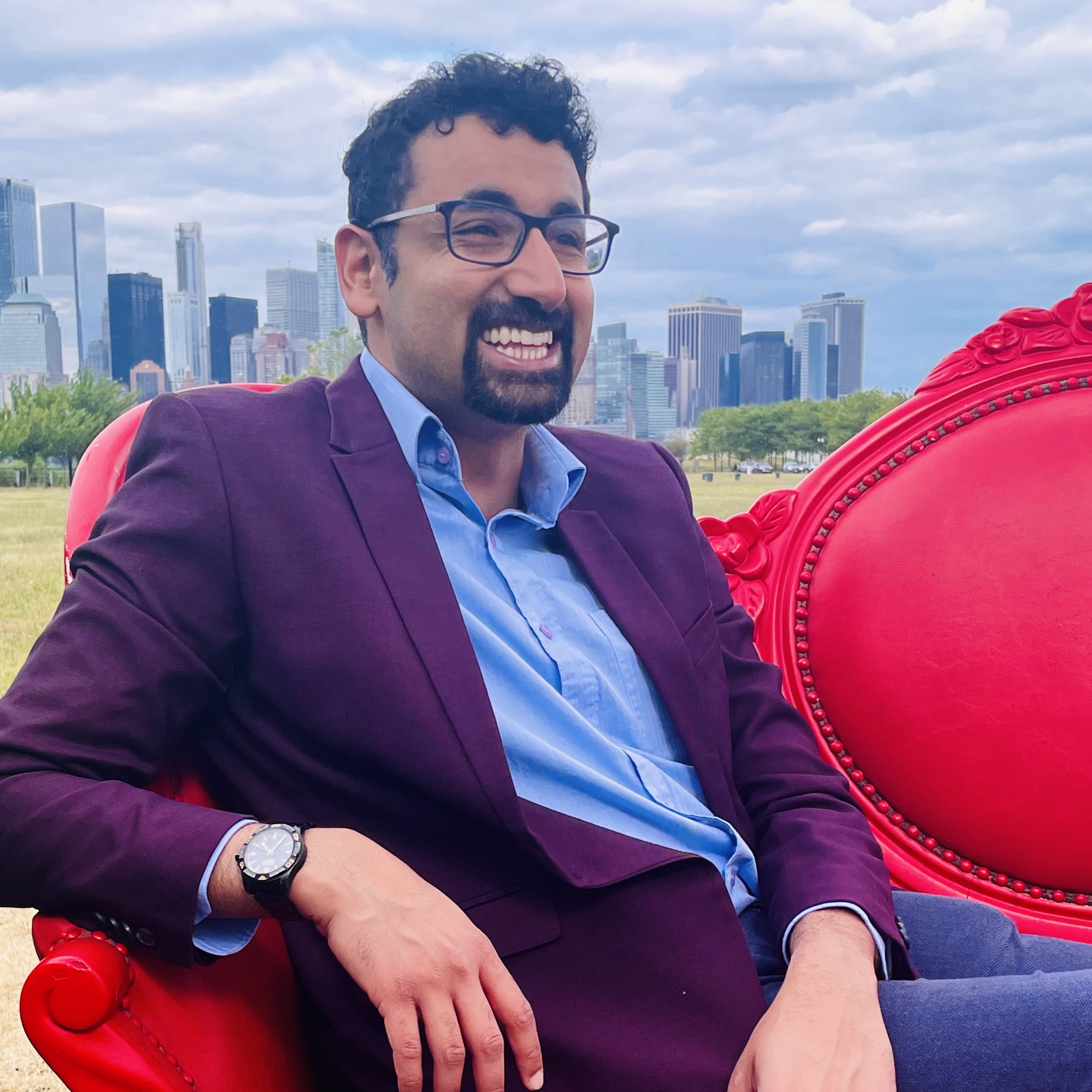 Sahil Luthra Person in a maroon blazer and blue shirt, laughing while seated on a red armchair in a park with the Chicago skyline in the background.