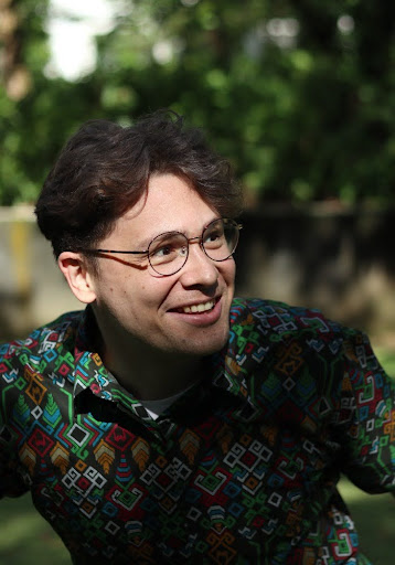 Maximilian Tegtmeyer Person in a vibrant geometric-patterned shirt and round glasses smiling outdoors with a blurred green background.