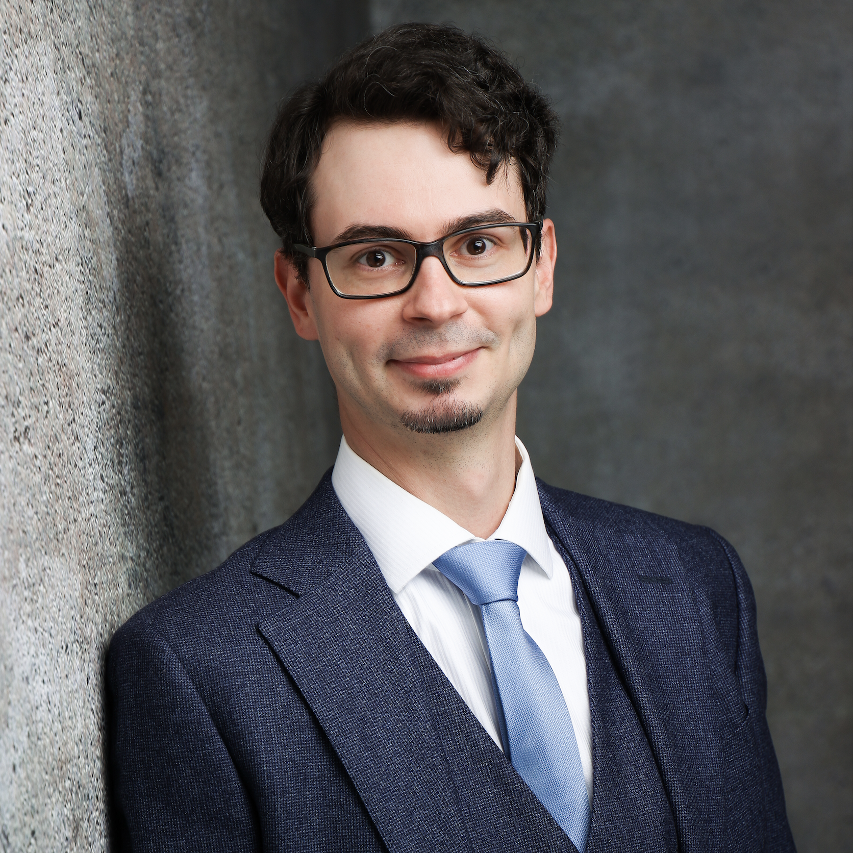 Jonathan Becker Portrait of a smiling individual in a formal blue suit and light blue tie, standing against a textured gray background.