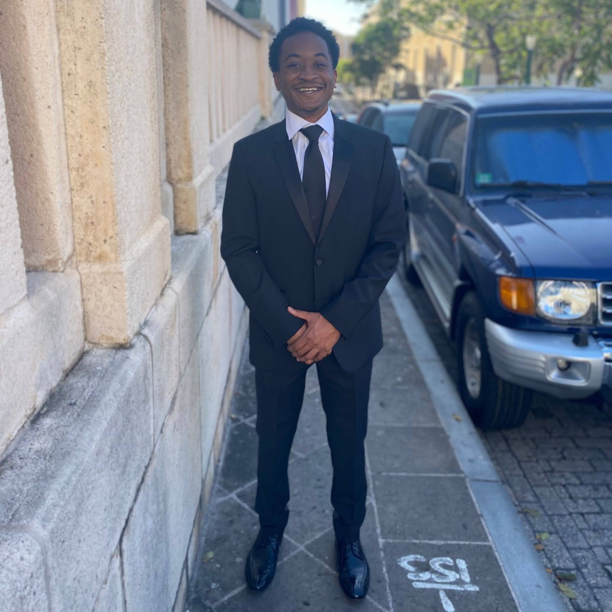 Raymond Blackwell Person in a black suit smiling while standing on a sidewalk next to a stone wall.