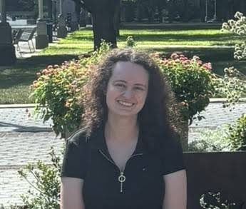 Rebecca Pearce Person smiling, seated outdoors with green plants and trees in the background.