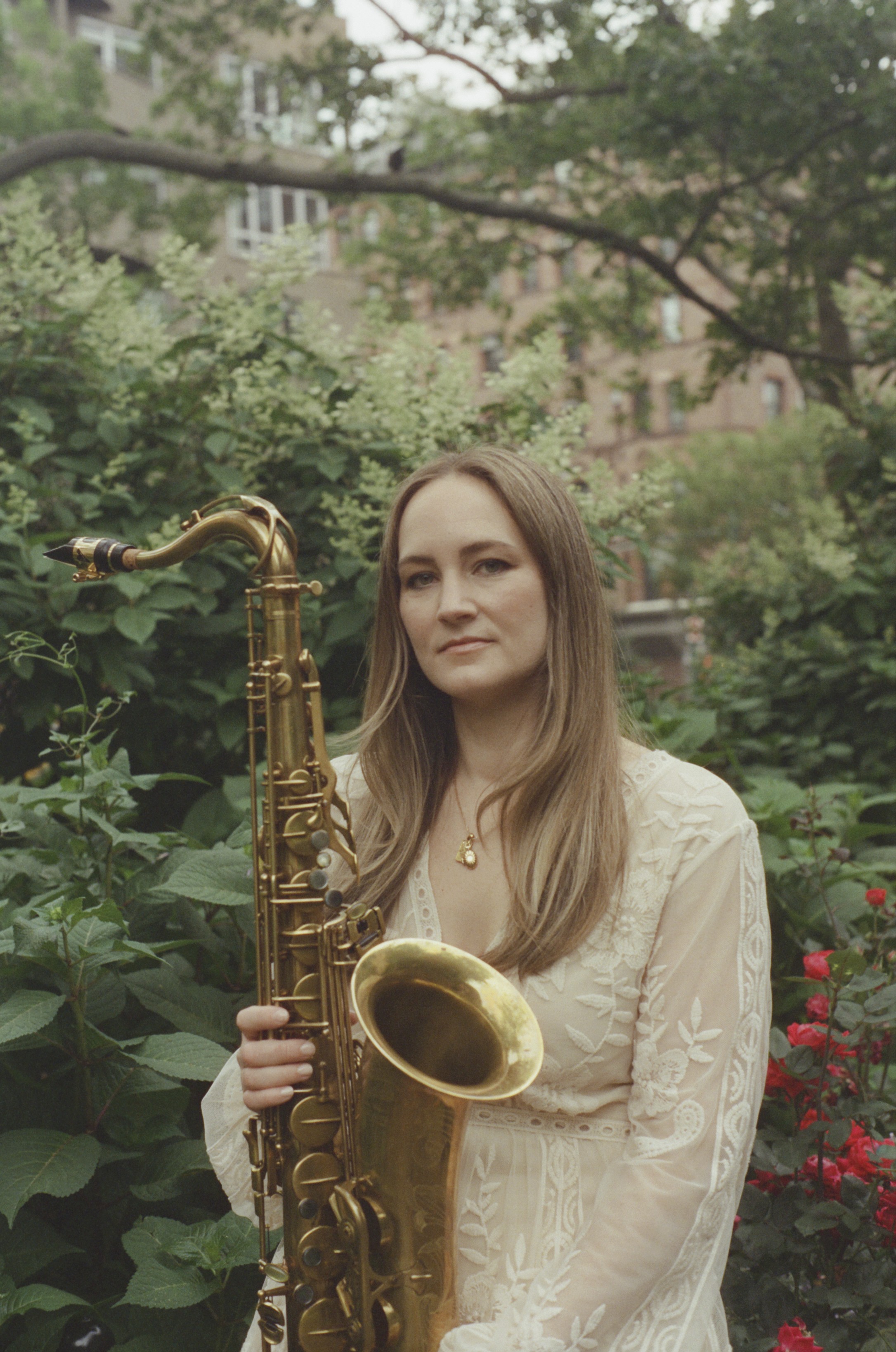 Roxy Coss Individual holding a saxophone stands amidst lush greenery with red flowers and a brick building visible in the background.