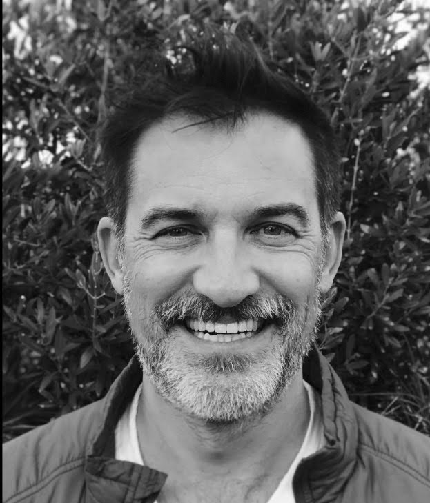 Scott Burkhardt Close-up portrait of a smiling person with short hair and a beard, standing in front of a leafy backdrop.