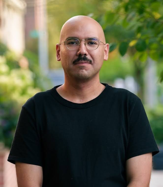 Sebastian Lopez Vergara Portrait of an individual with a shaved head wearing a black T-shirt, standing outdoors with greenery in the background.
