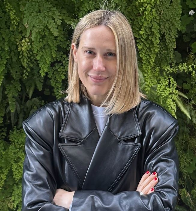 Valeria Meiller Person in a black leather jacket standing with arms crossed in front of a lush green bush.