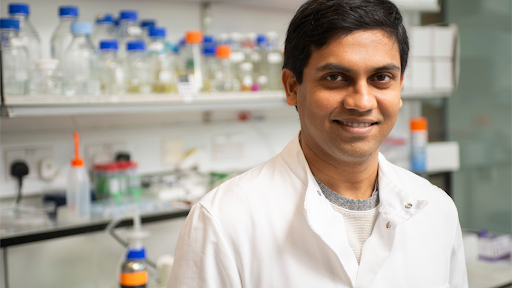 Viswanathan Chandrasekaran Scientist in a lab coat smiling at the camera in a laboratory filled with chemical bottles and lab equipment.