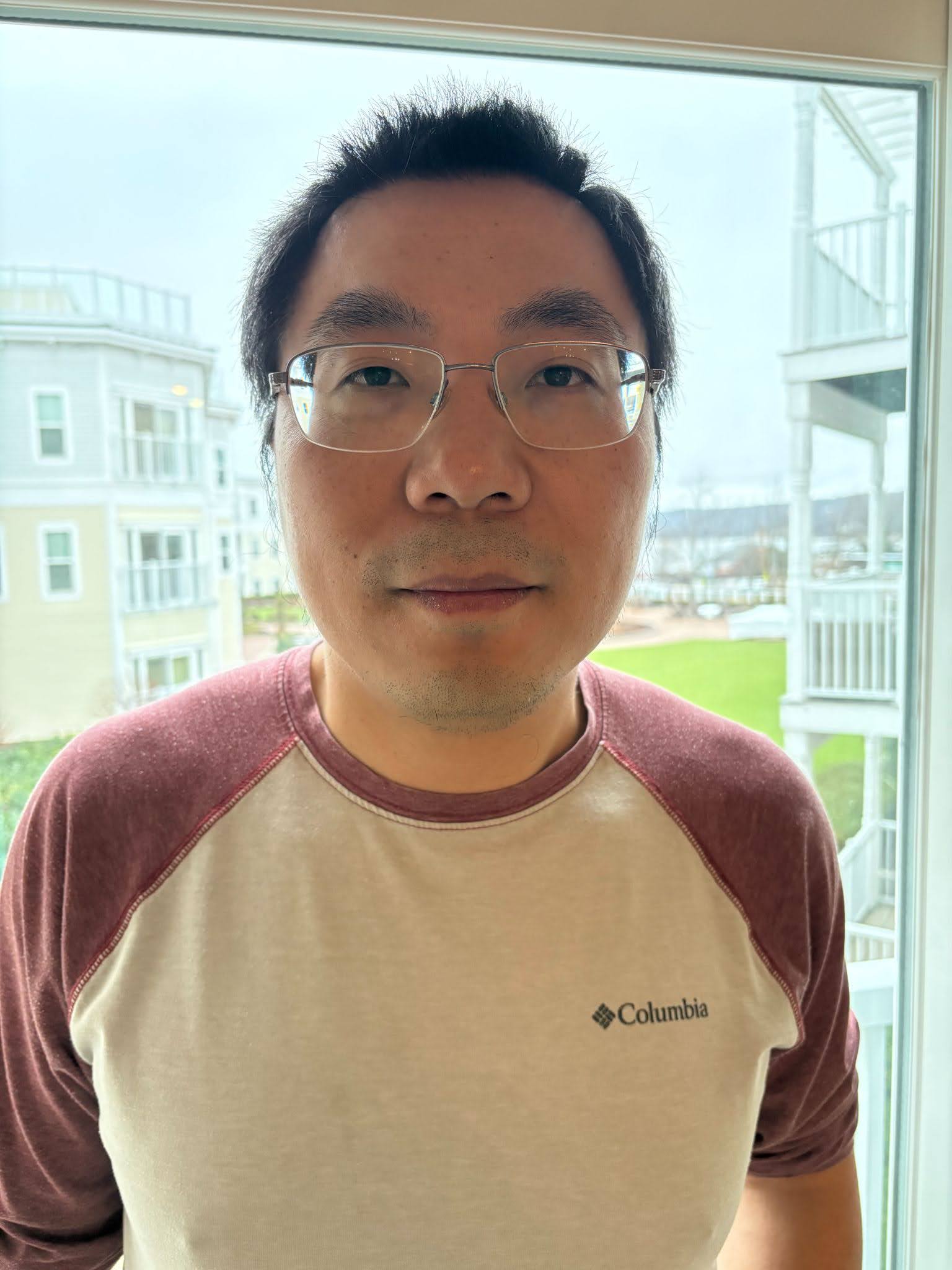 Yin-Chen He  Person wearing glasses and a Columbia branded t-shirt, standing in front of a window with a view of buildings and stairs outside.