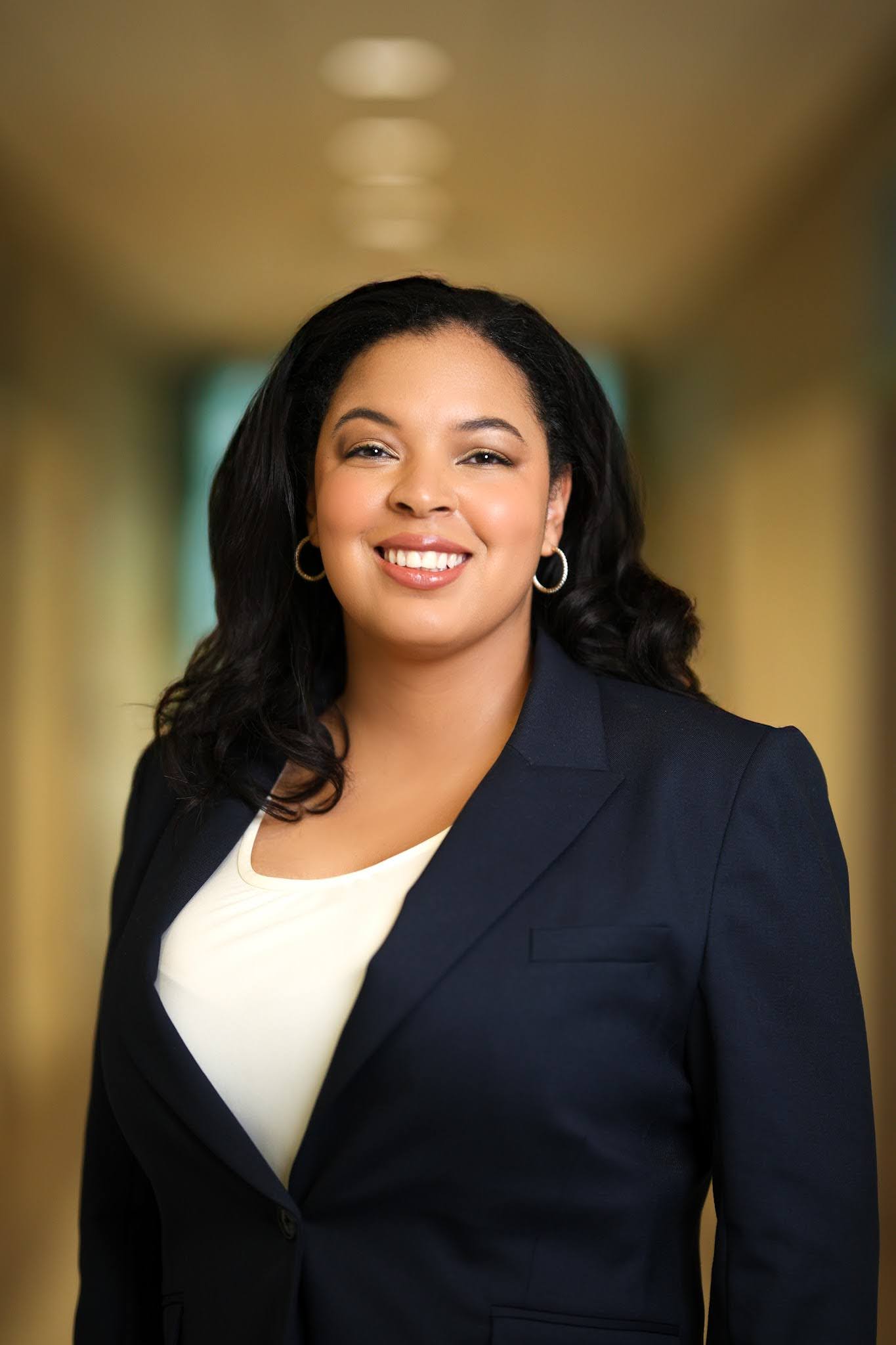 Yolanda Mackey-Barkers Professional portrait of a person smiling, wearing a dark suit and a light shirt, standing in a warmly lit corridor.