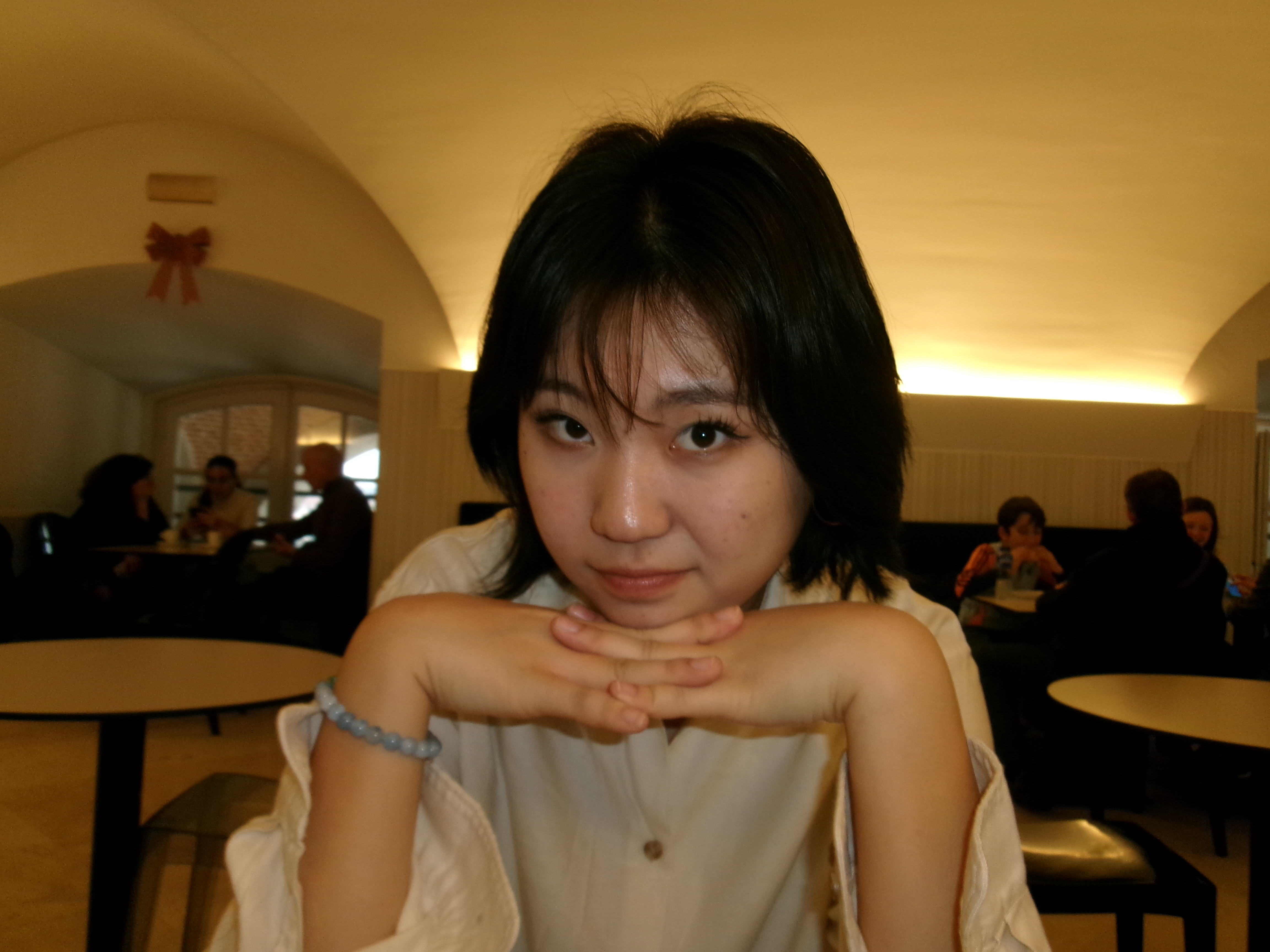 A person with short dark hair, in a white shirt, rests their chin on folded hands, smiling softly in a warmly lit cafe with people in the background.