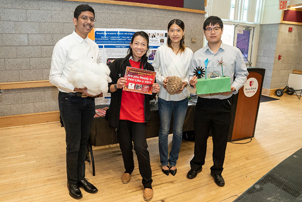 Four people stand in a room, smiling, each holding objects like cotton and a model. They're near a poster and a podium, creating a collaborative and positive atmosphere.