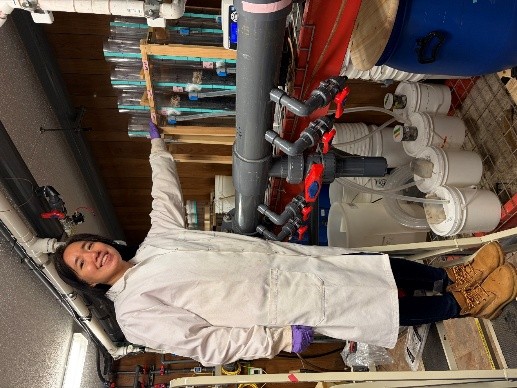 Xiumei Jian in a white lab coat, work boots, and purple gloves stands on a step stool in a water research facility, smiling as she rests her hand on a rack of tall, clear filtration columns. The laboratory environment is filled with industrial piping, red-handled valves, and various collection buckets and drums, suggesting an active experiment in water treatment or environmental engineering.