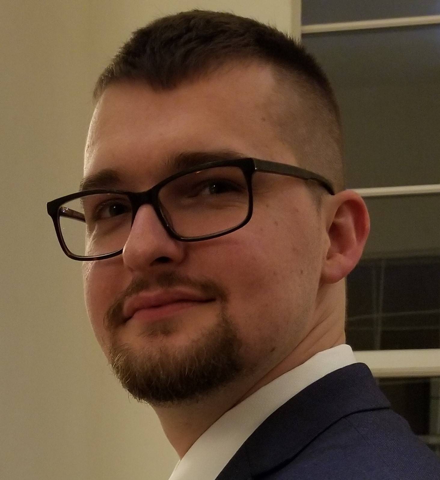 A close-up, chest-up headshot of a young man with light skin, short-cropped dark hair, and a neatly trimmed goatee. He is wearing dark-rimmed rectangular glasses and is angled slightly to his right, looking toward the camera with a subtle smile. He is dressed professionally in a white collared shirt and a dark blue suit jacket. The background is a simple, neutral-toned wall with a hint of a window frame or architectural detail to the right.
