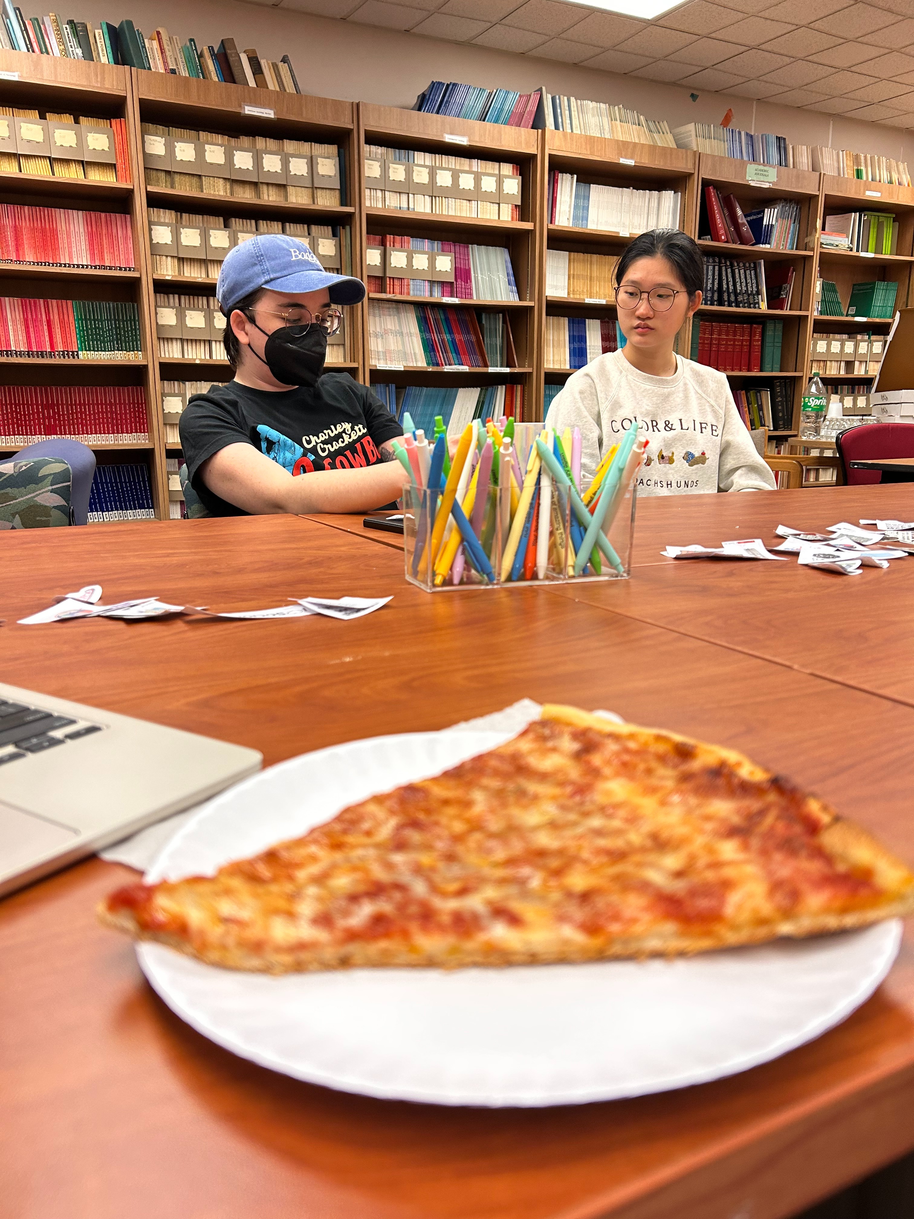 People sitting at a table in a library with bookshelves, a container of colorful pens, and a slice of pizza on a plate.